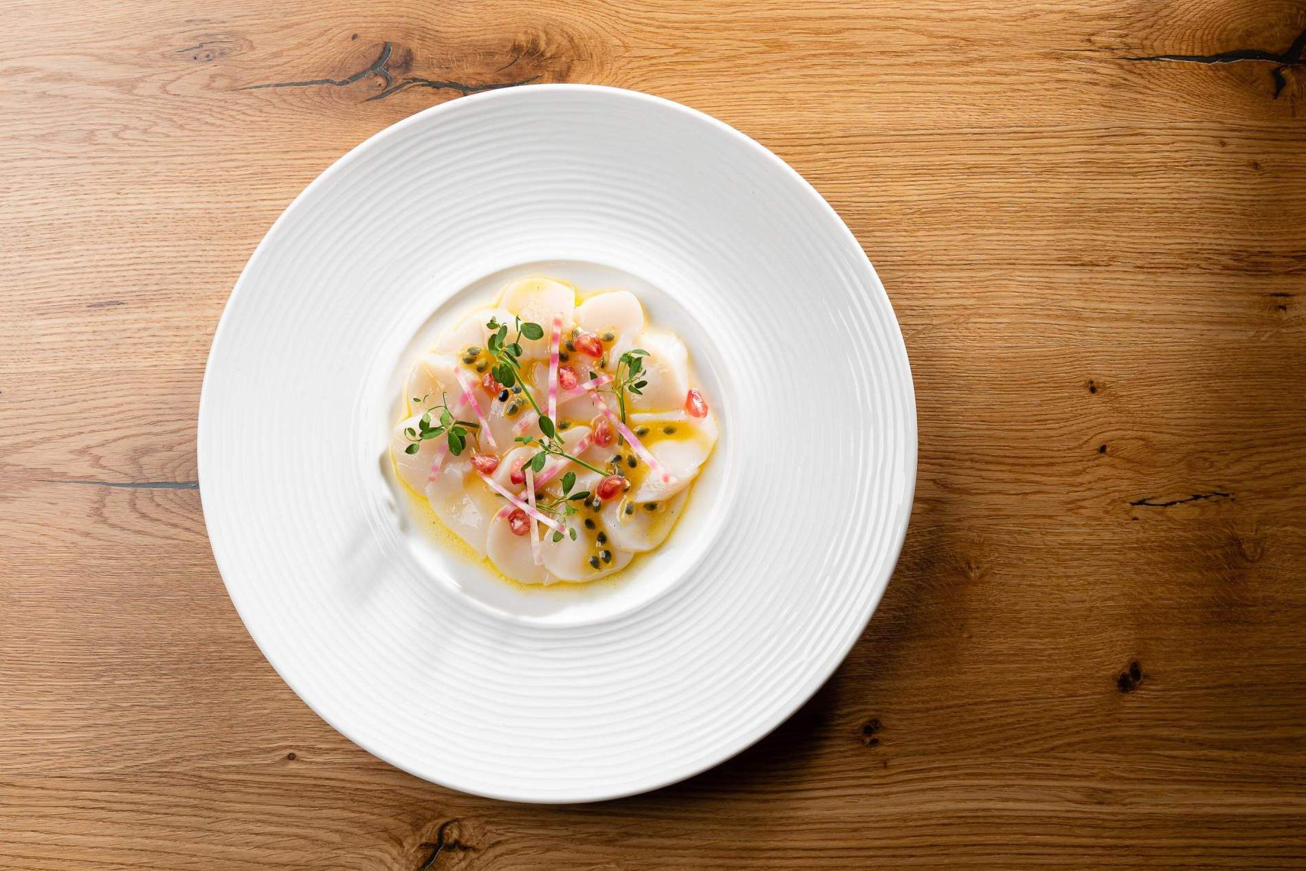 Carpaccio de Saint-Jacques, fruit de la passion, grenade et radis rose, servi sur une table en chêne